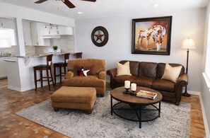 Un salon avec un canapé en cuir marron, un pouf assorti et une table basse ornée de bougies et de livres. Un tableau représentant des chevaux est accroché au mur, et une décoration en forme d’étoile est placée au-dessus de la cheminée.