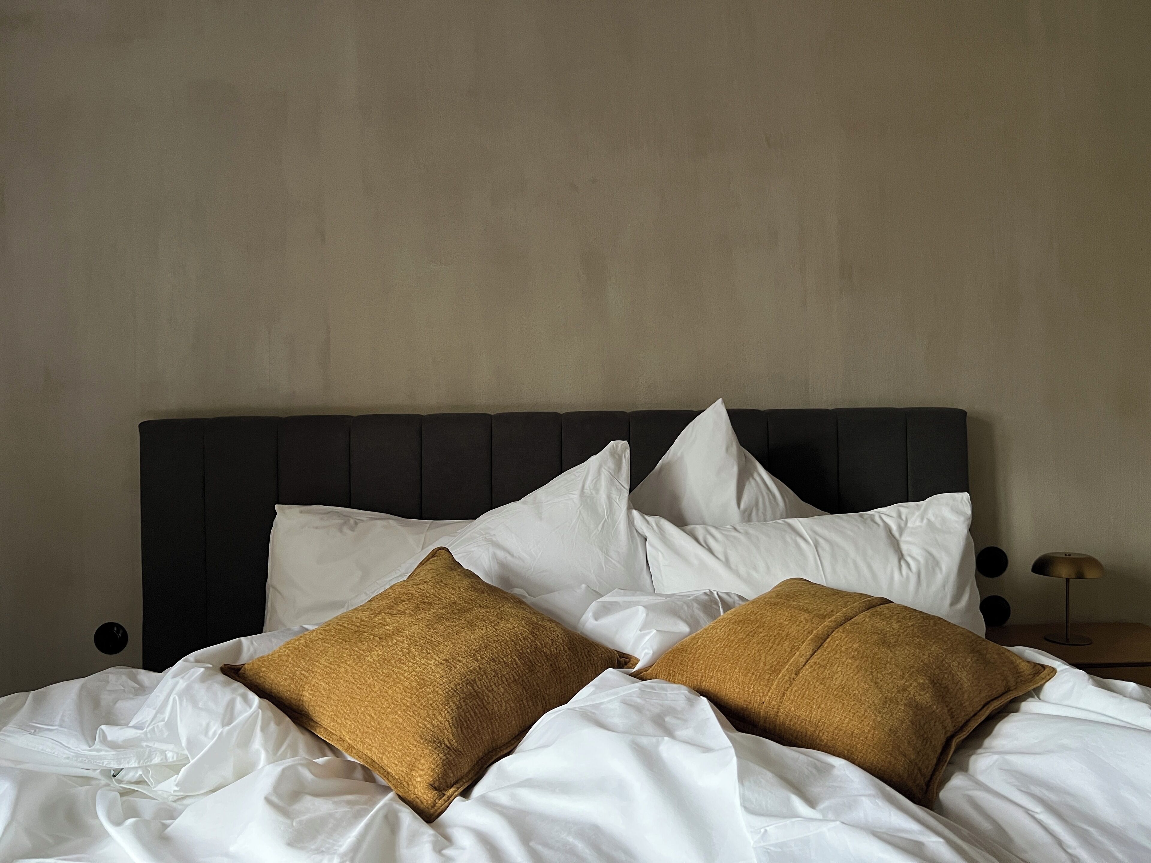 Een bed met witte lakens en twee bruine kussens, een donker hoofdbord en een effen muur.