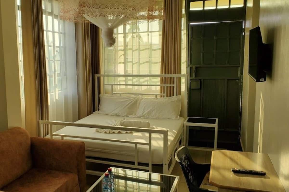 Een eenpersoonskamer met een bed met wit metalen frame, een klein nachtkastje, een stoel en een aan de muur bevestigde televisie.