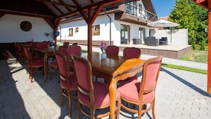 Ein Essbereich im Freien mit Holztisch und -stühlen, einer überdachten Terrasse und Blick auf ein Haus mit Balkon.