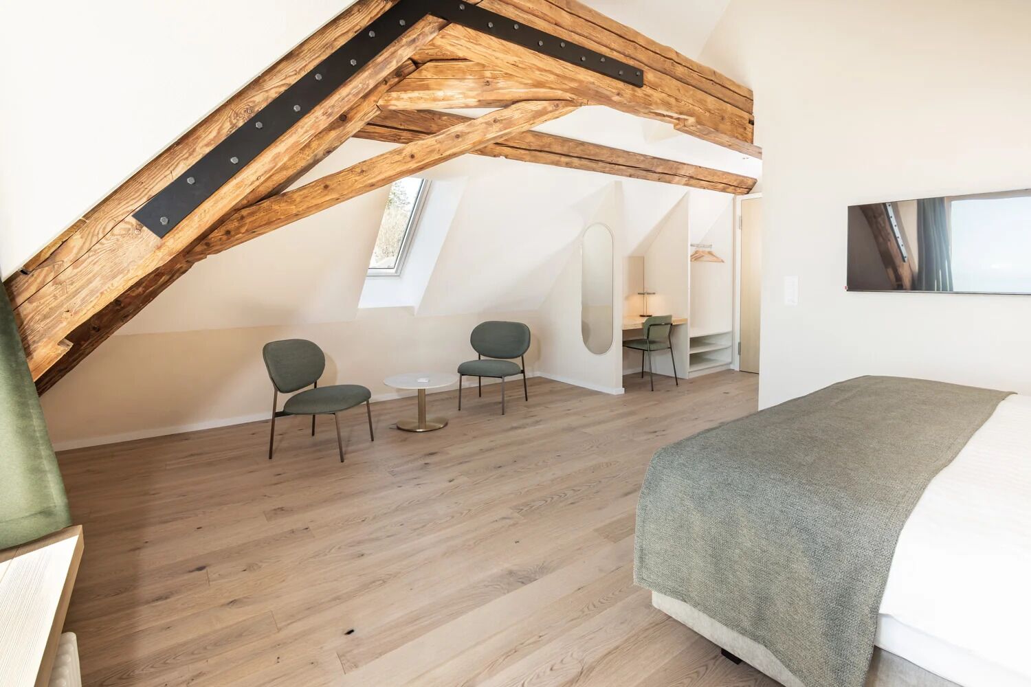 Een slaapkamer in loftstijl met zichtbare houten balken, een bed, twee stoelen, een tafeltje en een spiegel.