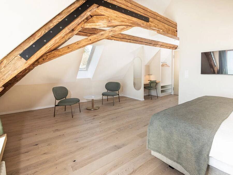 Een slaapkamer in loftstijl met zichtbare houten balken, een bed, twee stoelen, een tafeltje en een spiegel.