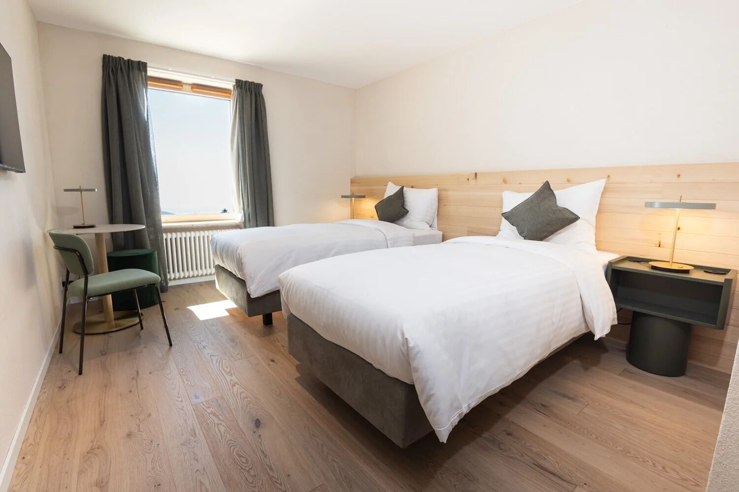 A hotel room with two beds, a small table, a chair, a nightstand, and a window with curtains.