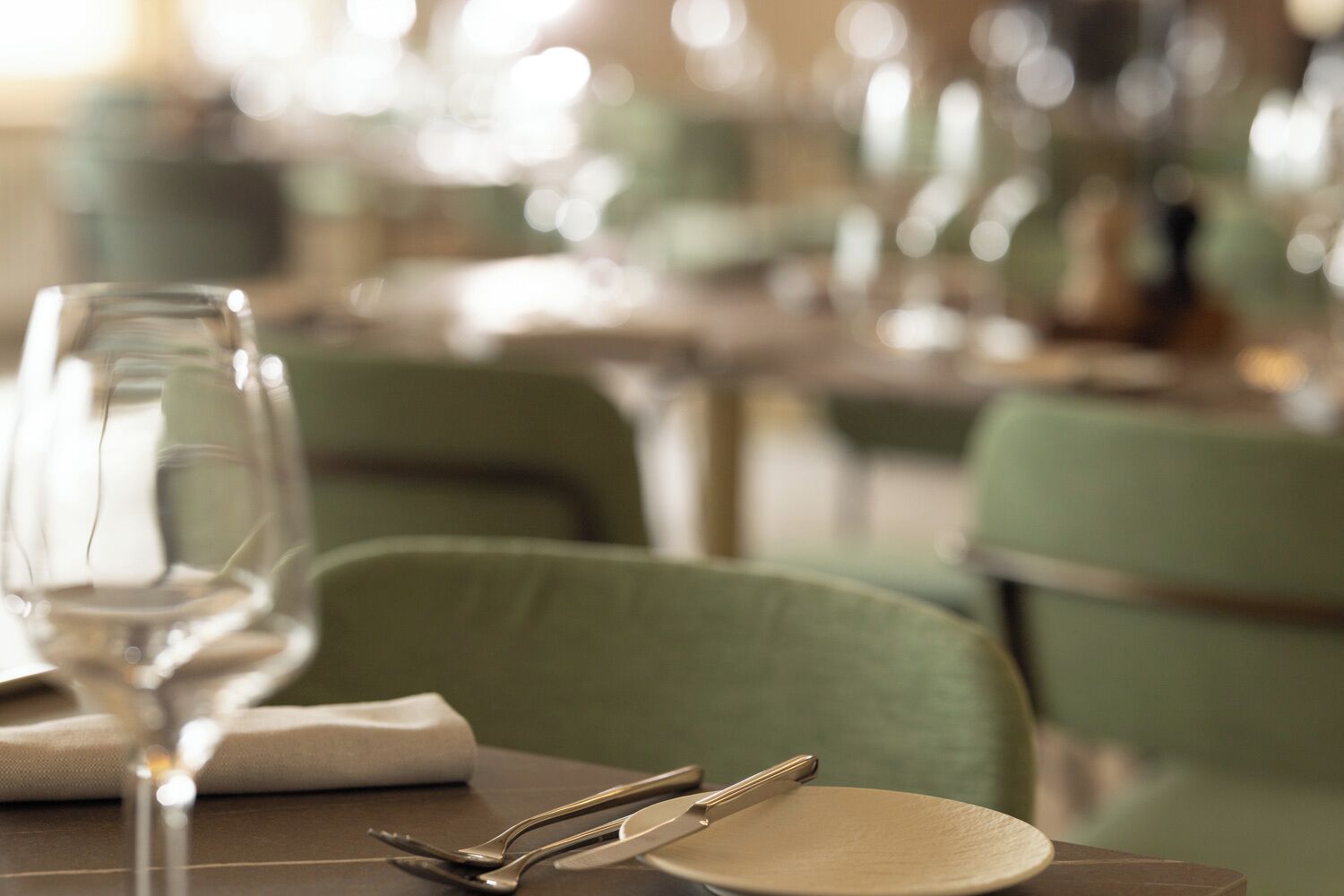 Ein Esstisch mit einem Weinglas, Besteck und einem Teller, in einem Restaurant mit grünen Stühlen und unscharfem Hintergrund.