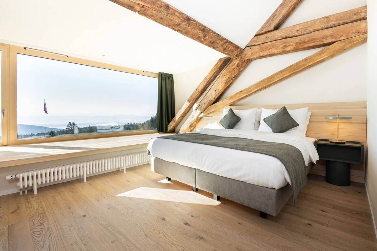 Une chambre avec une grande fenêtre, un lit avec des oreillers, une table de chevet et un radiateur.
