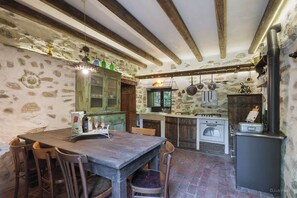 Uma cozinha rústica com paredes de pedra, vigas de madeira, uma grande mesa de madeira e um fogão antigo.