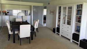 Een eethoek met een tafel en stoelen, een witte boekenkast, en een keuken op de achtergrond.