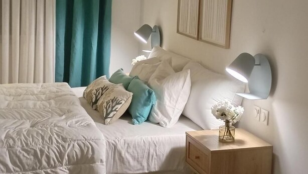 Una cama bien hecha con ropa de cama blanca y almohadas decorativas, una mesita de noche con lámpara y un aire acondicionado de pared.