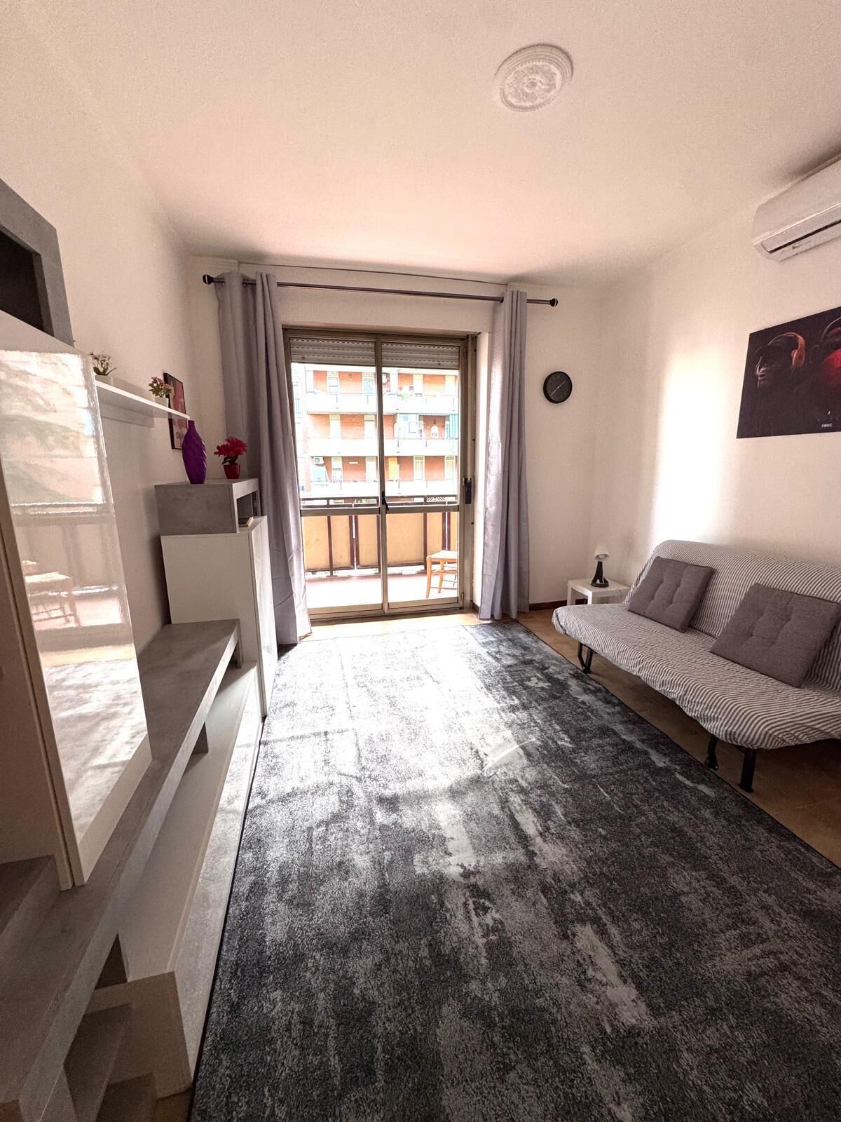 A modern living room with a sofa, a small table, a TV, and a balcony with a view of buildings.