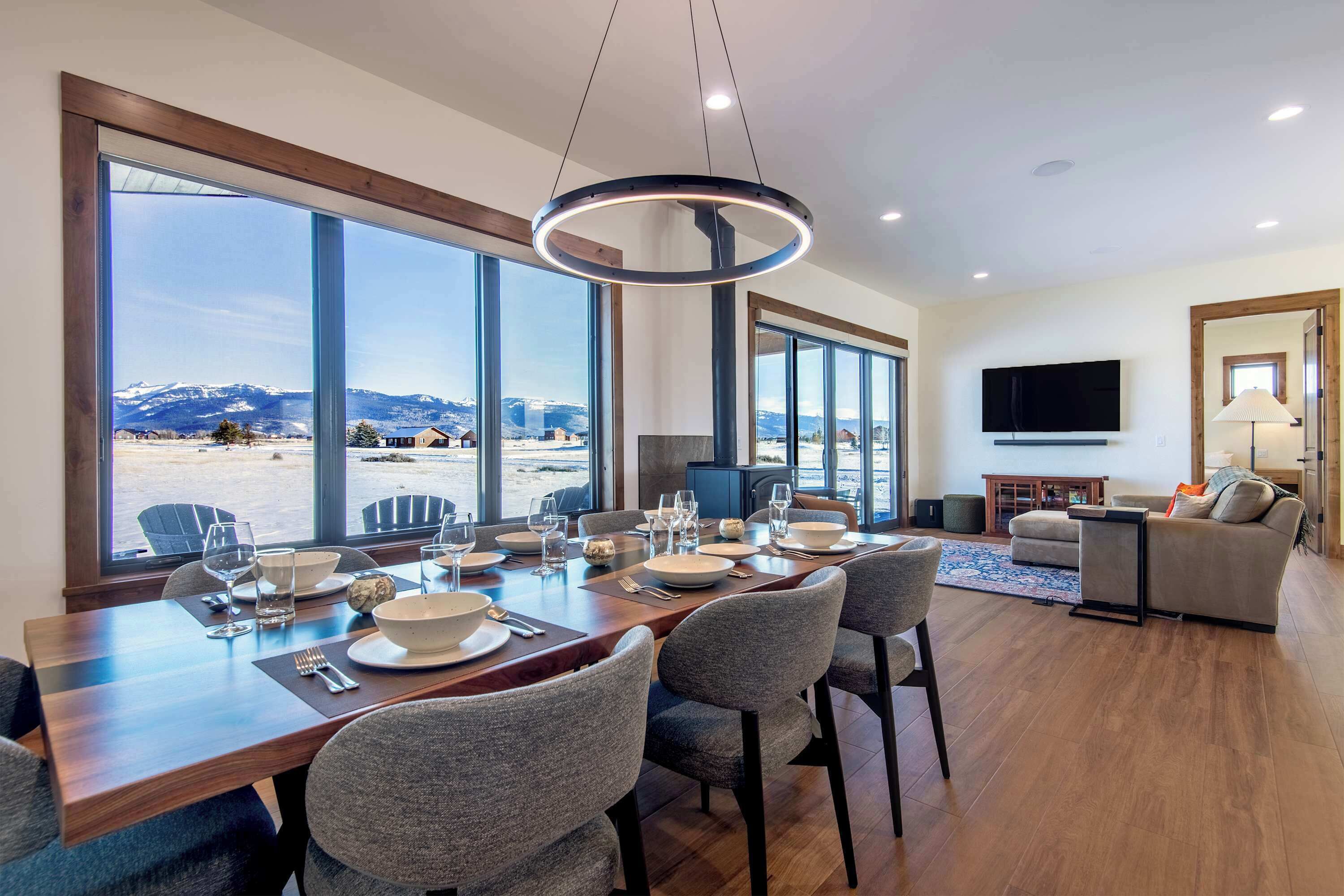A dining area with a long table set for four, large windows offering a mountain view, a flat-screen TV mounted on the wall, and a cozy seating area with a sofa and armchairs.