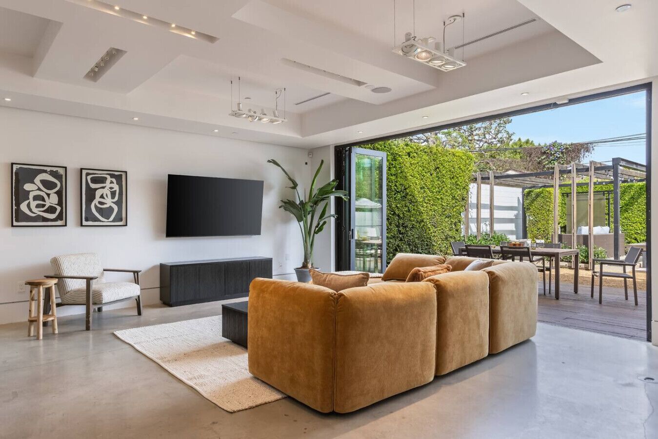 Un salon moderne doté d’un grand canapé d’angle, d’un téléviseur à écran plat, d’œuvres d’art abstraites accrochées au mur et offrant une vue sur l’extérieur grâce à de grandes portes vitrées.