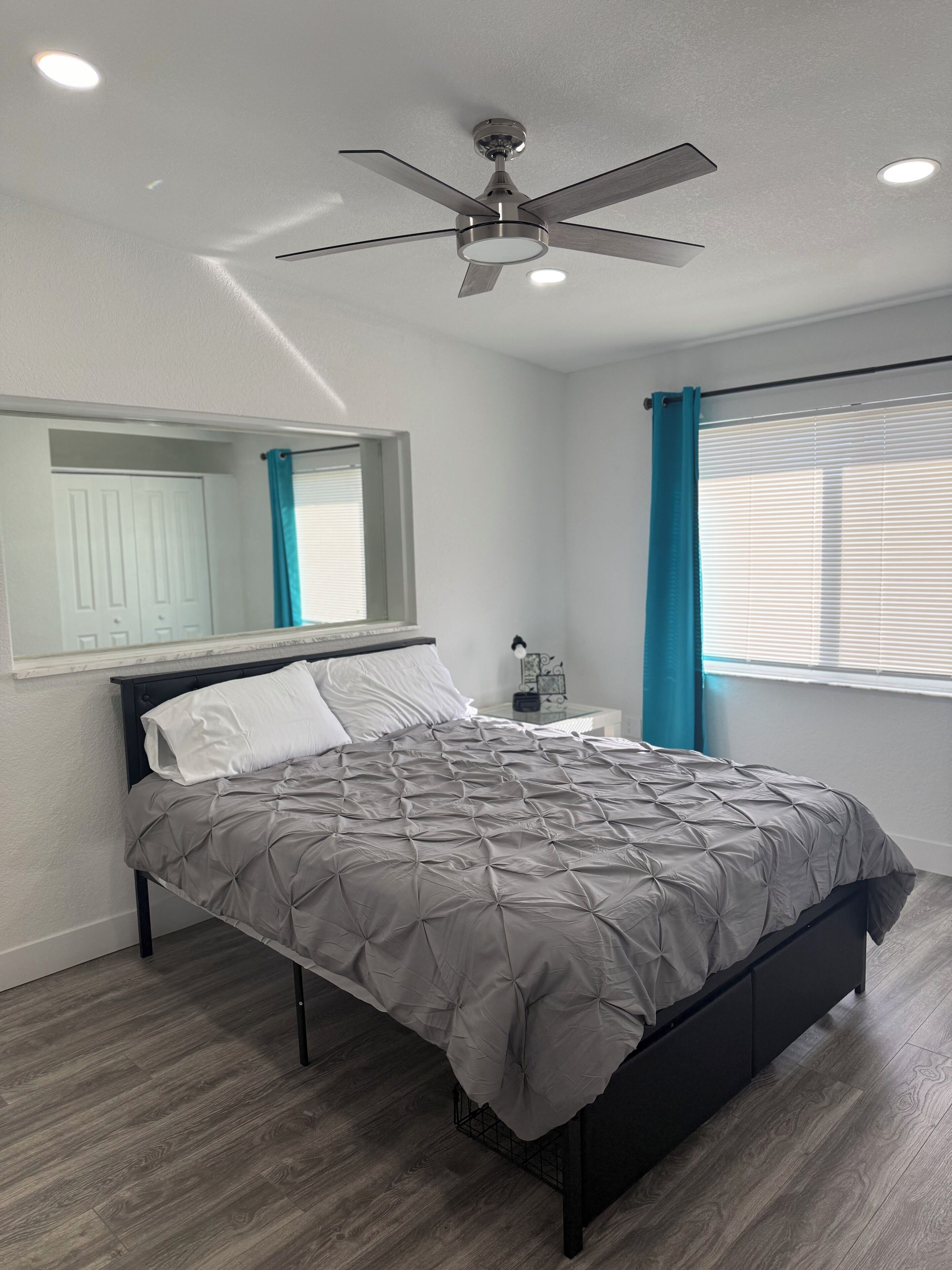 Une chambre à coucher moderne avec un lit, un ventilateur de plafond, une fenêtre avec des rideaux et une table de chevet avec une lampe.
