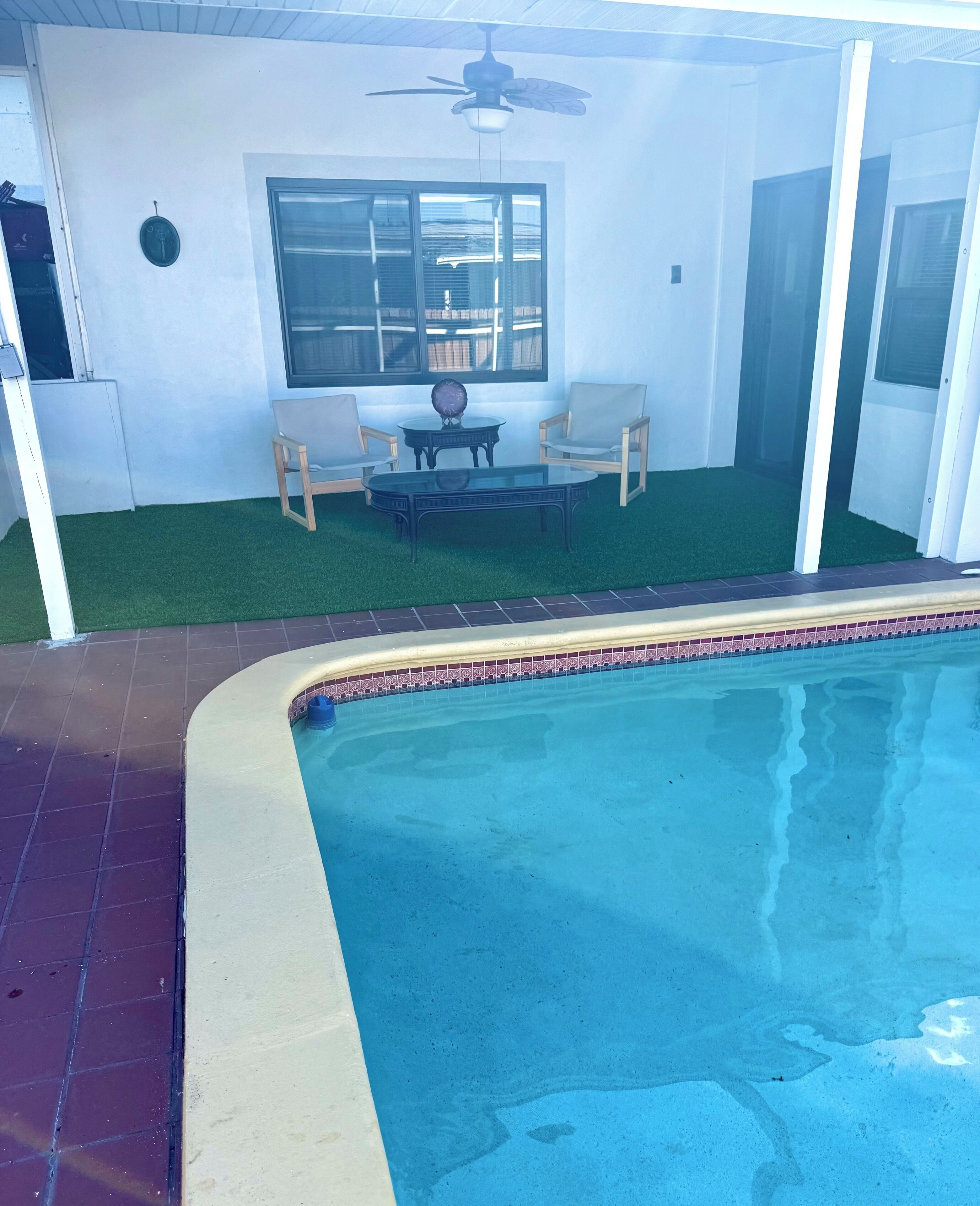 A swimming pool with a tiled edge, a ceiling fan, and a seating area with two chairs and a table.