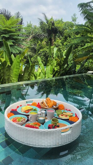 Ein Frühstücksbuffet mit verschiedenen Speisen und Getränken an einem Glastisch am Pool, umgeben von üppigem Grün.