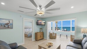 Ein geräumiges Wohnzimmer mit Deckenventilator, Fernseher, Esstisch und Meerblick durch große Fenster.