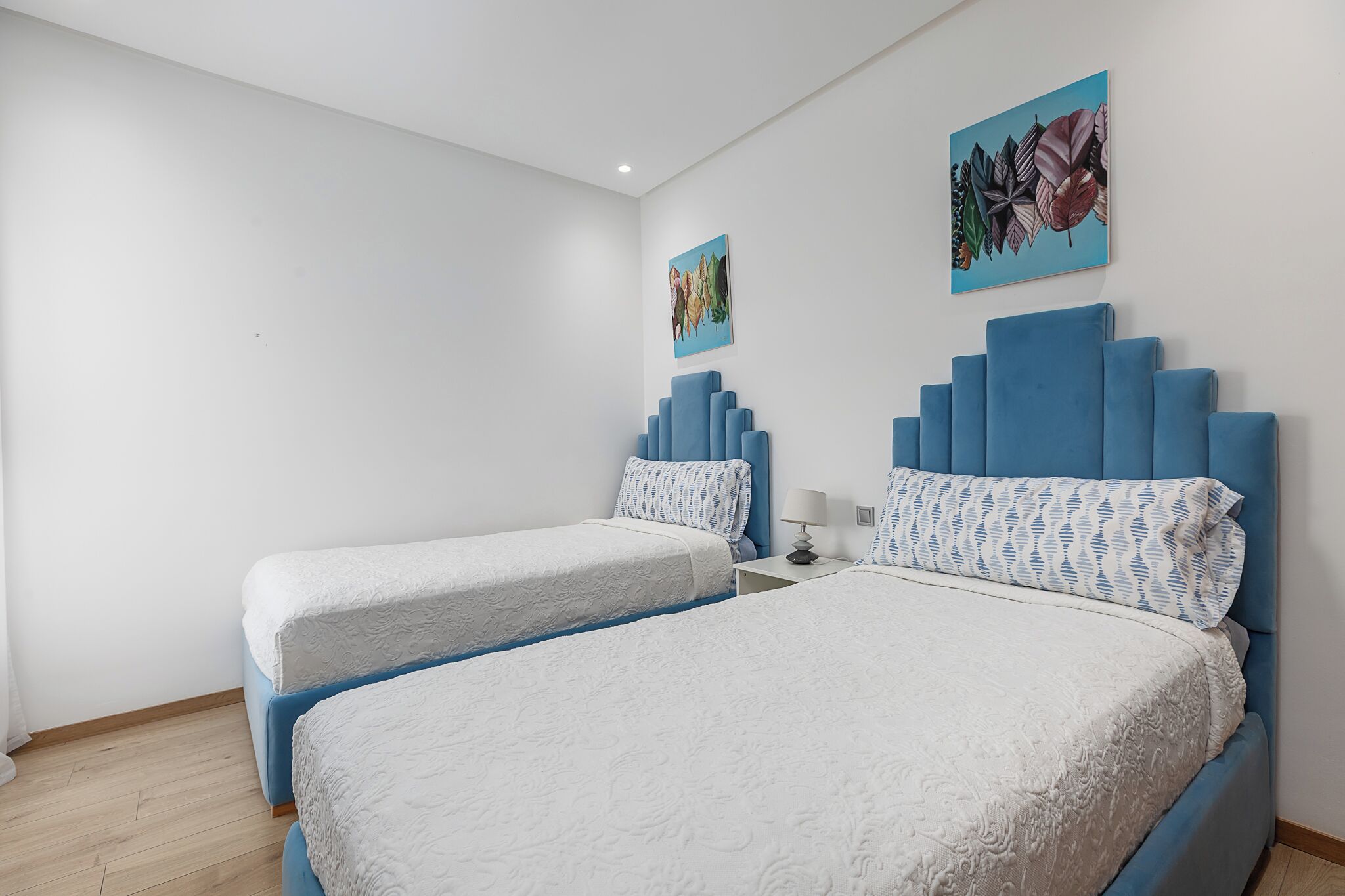 A hotel room with two beds, a blue headboard, a white wall, and a painting.