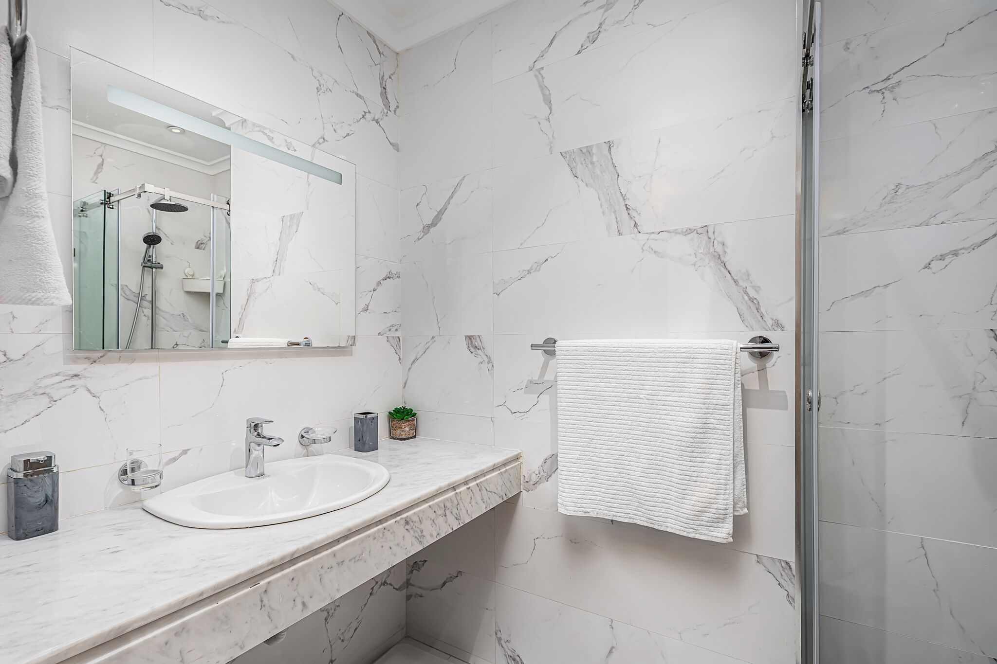 A modern bathroom with a marble sink, a glass shower enclosure, and a white towel hanging on the wall.
