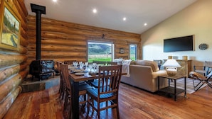 Interior de uma cabana de tronco com uma mesa de jantar, cadeiras, um sofá, uma televisão e uma lareira.
