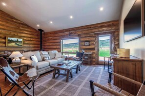 A log cabin interior with a sofa, armchair, table, and a view through large windows. - Teton Homestead: Log Cabin on 4 Acres w/ Hot Tub (Driggs)