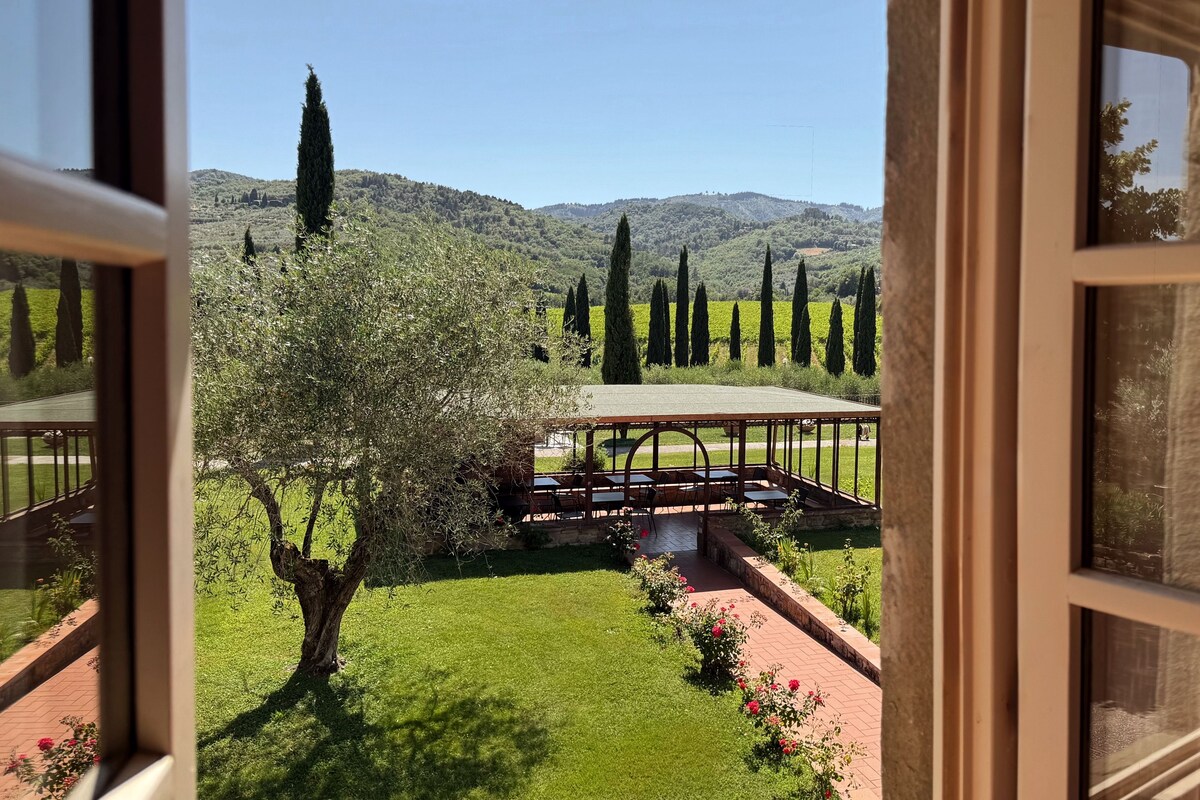 En vy genom ett fönster ut mot en trädgård med en pergola, ett träd och ett avlägset kullområde.