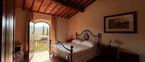 A bedroom with a bed, a nightstand, a lamp, a framed picture on the wall, and a view through a window.