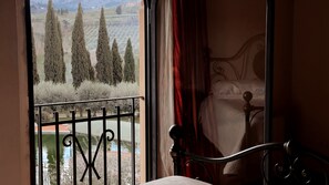 A room with a bed, a window with a view of trees and a distant building, and a balcony with a railing.