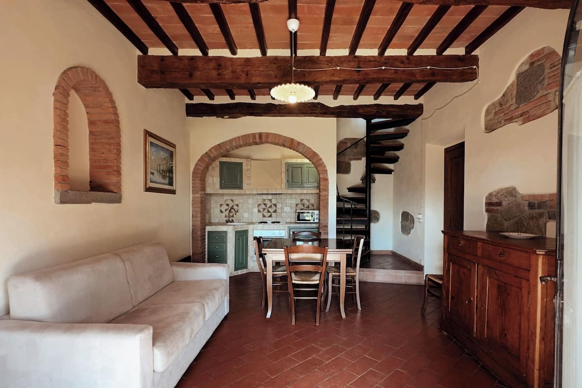 A living room with a sofa, dining table, chairs, a kitchen with a tiled backsplash, and a staircase.