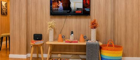 Habitación con televisor de pantalla plana montado en la pared, un escritorio de madera con una computadora portátil, un taburete pequeño y objetos decorativos como jarrones y una cesta tejida de colores.