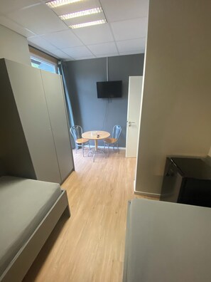 A room with a round table, four chairs, a television mounted on the wall, and a door. - Bordinghouse Halverde (Hopsten)