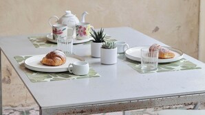 Ein Tisch mit einem Croissant, einem Teekessel, einer Tasse, einem Untertasse, einer kleinen Topfpflanze und einem Glas Wasser.