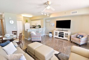 A living room with a sofa, ottoman, and two patterned armchairs. There is a dining table with chairs, a kitchen area, and a TV mounted on the wall. - ☀️Stunning Beach Front | Large Pool | Shores of Panama 530☀️ (Panama City Beach)