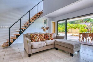 A modern living room with a beige sectional sofa, a wooden staircase, and a glass door leading to an outdoor area with a table and chairs.