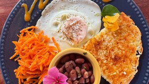 A plate with a fried egg, hash browns, shredded carrots, kidney beans, and sliced cucumbers.