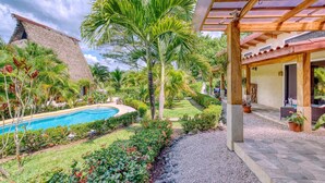 A pool surrounded by lush greenery, a thatched-roof building, and a covered patio area.