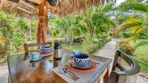 An outdoor dining area with a wooden table set for two, surrounded by lush tropical vegetation.