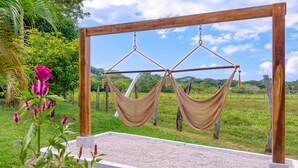 A hammock swing set with two hammocks suspended from a wooden frame, set against a grassy field with trees and a clear sky.