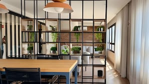 Un espace repas moderne avec une table et des chaises en bois, une cloison en métal noir avec des panneaux de verre, et une vue sur la cuisine et l’espace de vie à travers le verre.