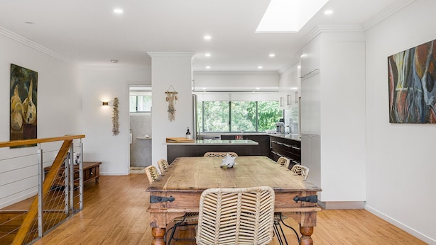 Un'ampia sala da pranzo con un grande tavolo in legno e sedie in vimini, una cucina con elettrodomestici moderni e vista sul verde attraverso ampie finestre.