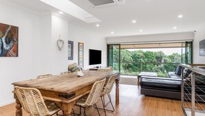 Un'ampia zona giorno con un tavolo da pranzo in legno, sedie in vimini, una televisione a schermo piatto e un balcone con vista sul verde.