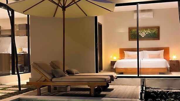 A modern hotel room with a large bed, a bedside table with a lamp, a small table with a chair, and a view of the pool area. - Marvelous 3-bedroom villa with AC in vibrant Ubud Bali (Bali)