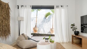 A modern living room with a large window, white curtains, a sofa, a lamp, and a TV on a wooden stand.