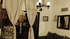 A cozy living room with a wooden table, dark sofa, and curtains. A clock and chandelier are visible on the walls.