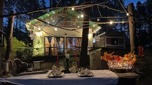 An outdoor dining area with a covered table set for two, surrounded by trees and a building in the background.