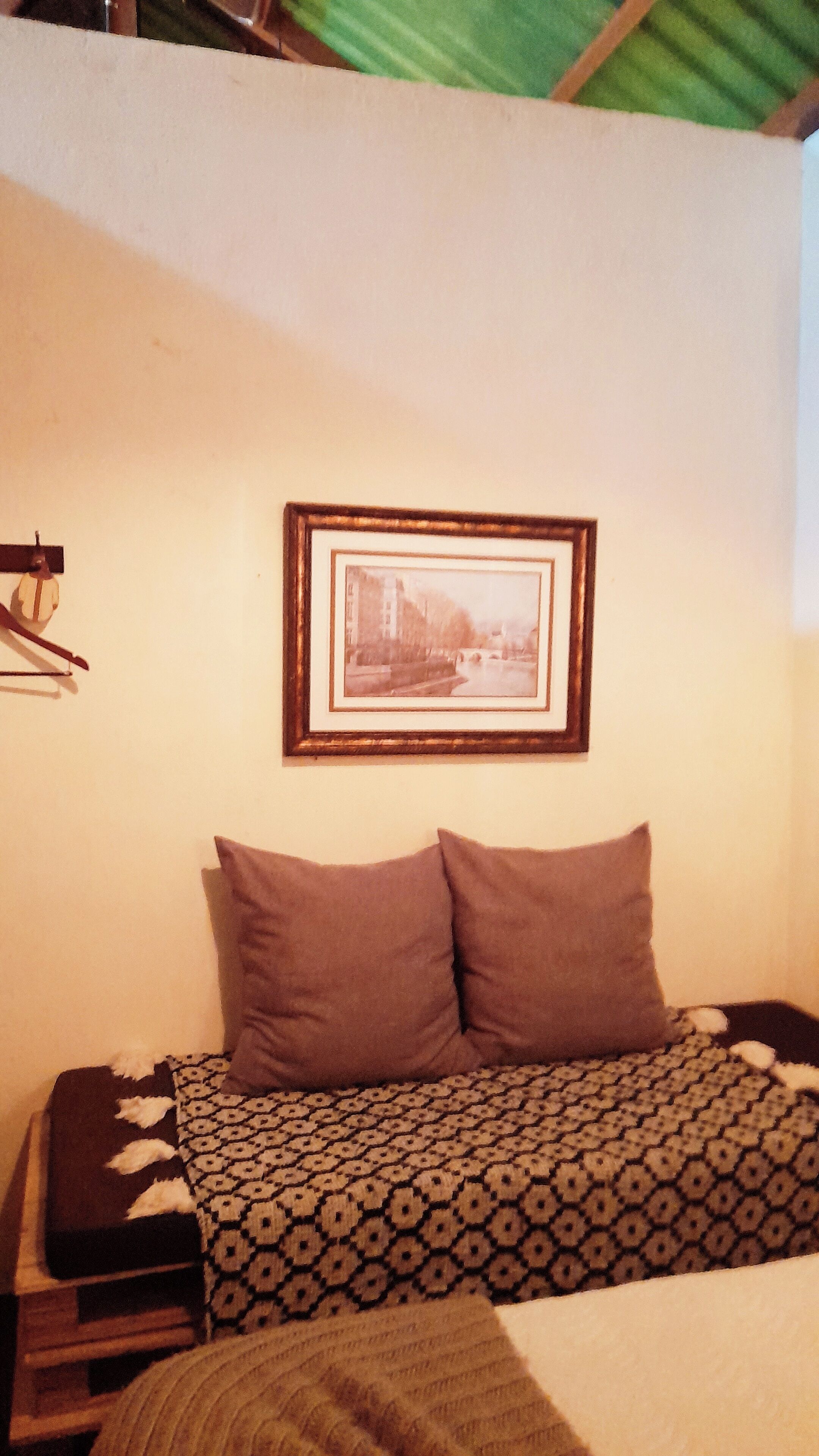 Een kamer met een bed, een bedekt dek, twee kussens, een ingelijst schilderij aan de muur en een kleine tafel met een lamp.