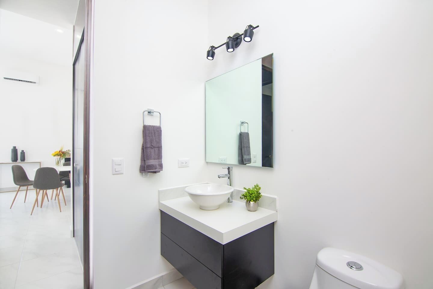 A modern bathroom with a white sink, black cabinet, and a large mirror. There is a small plant on the sink counter and a towel hanging on a rack.