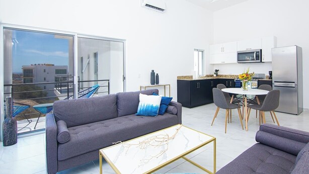 Un appartement moderne avec un canapé gris, une table à manger avec des chaises, un coin cuisine et un balcon avec vue.