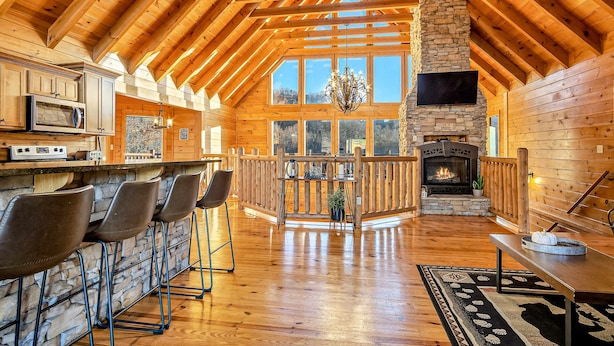 A spacious wooden cabin with a stone fireplace, a bar area with stools, and large windows offering a view of the surrounding landscape. - Luxe Sister Cabins, Sleeps 24, Pool, Hot Tub, Games (Gatlinburg)