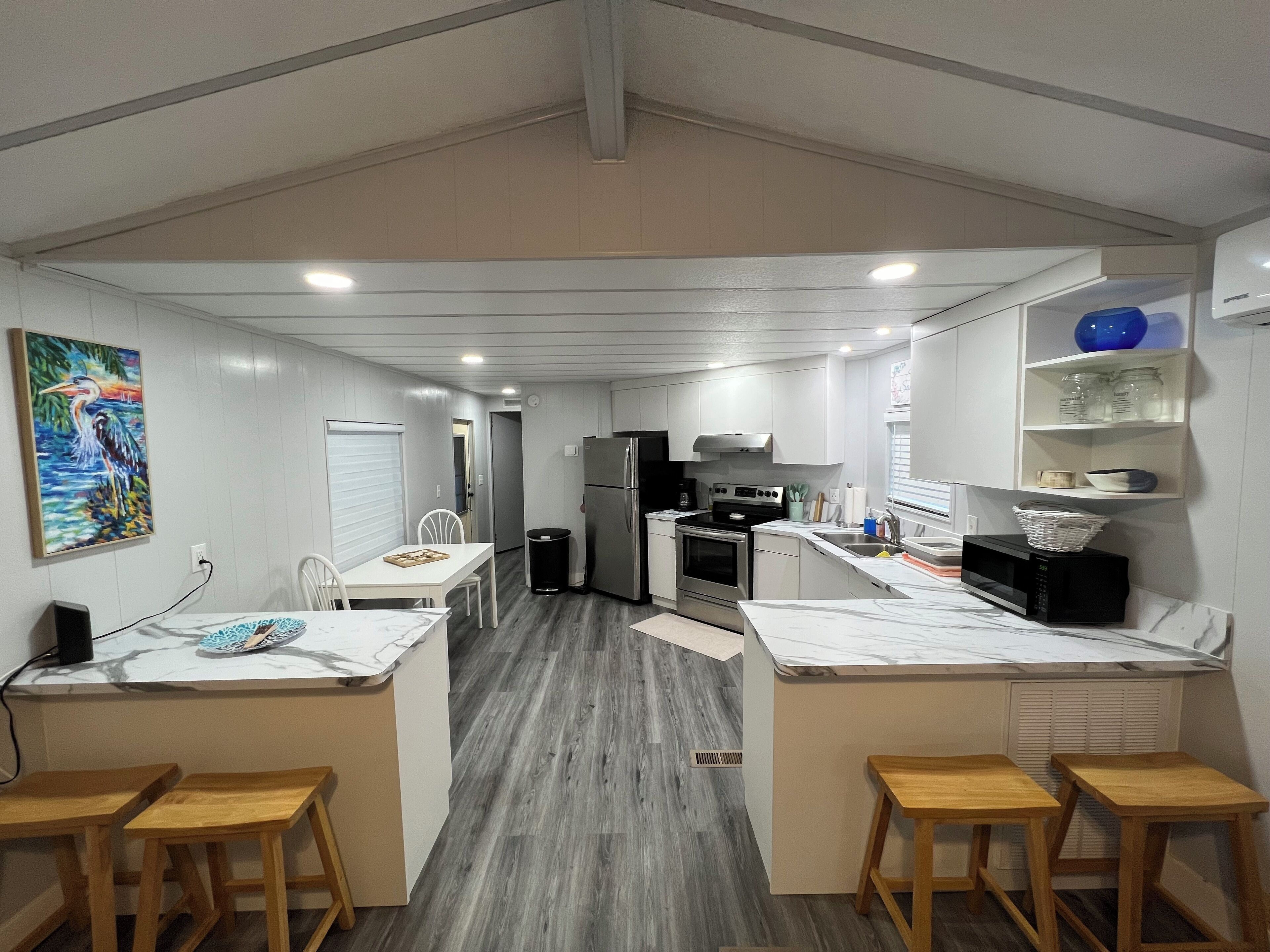A modern kitchen with white cabinets, a stainless steel refrigerator, a microwave, and a sink. There are wooden stools and a table with a white tablecloth.