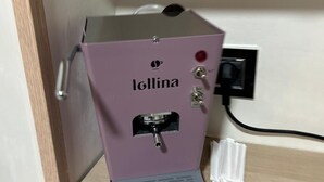 Una máquina de café con la marca 'Iollina' sobre una estantería en una habitación con una puerta de madera y una pared blanca.