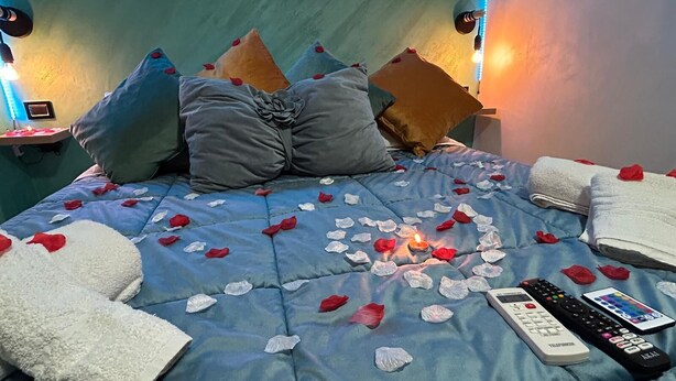 Una cama con pétalos de rosa, una vela encendida y dos mandos a distancia.